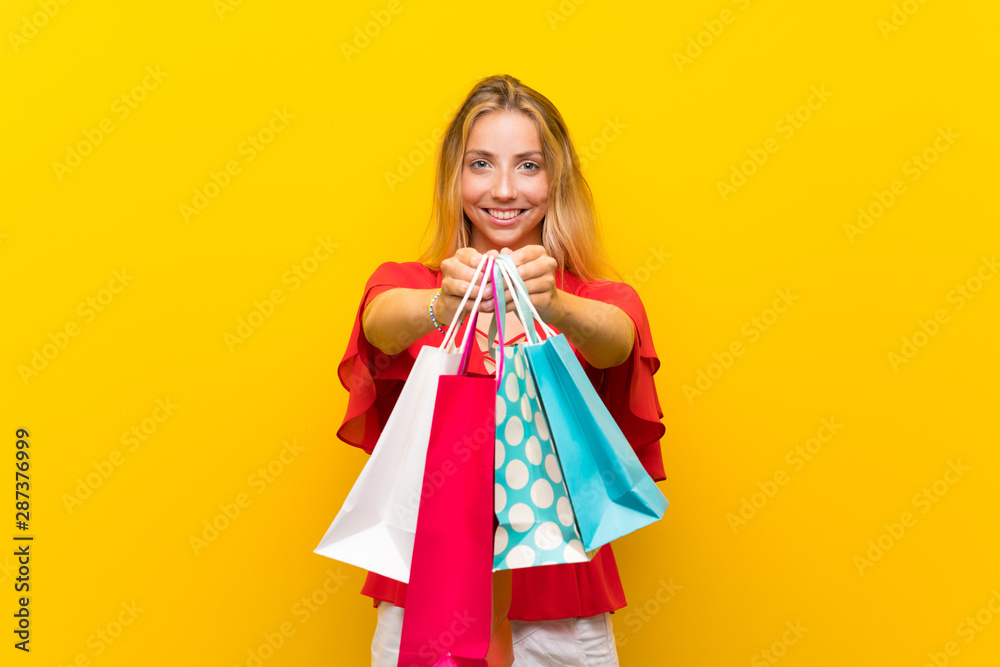 Blonde young woman over isolated yellow background holding a lot of shopping bags