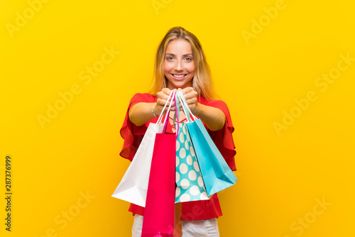 Blonde young woman over isolated yellow background holding a lot of shopping bags