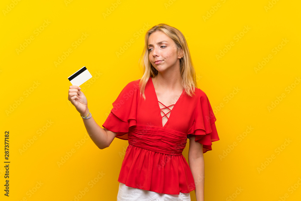 Blonde young woman over isolated yellow background holding a credit card Blonde young woman over isolated yellow background holding a credit card