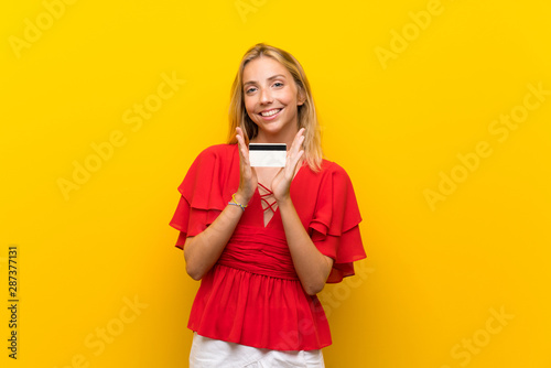 Blonde young woman over isolated yellow background holding a credit card