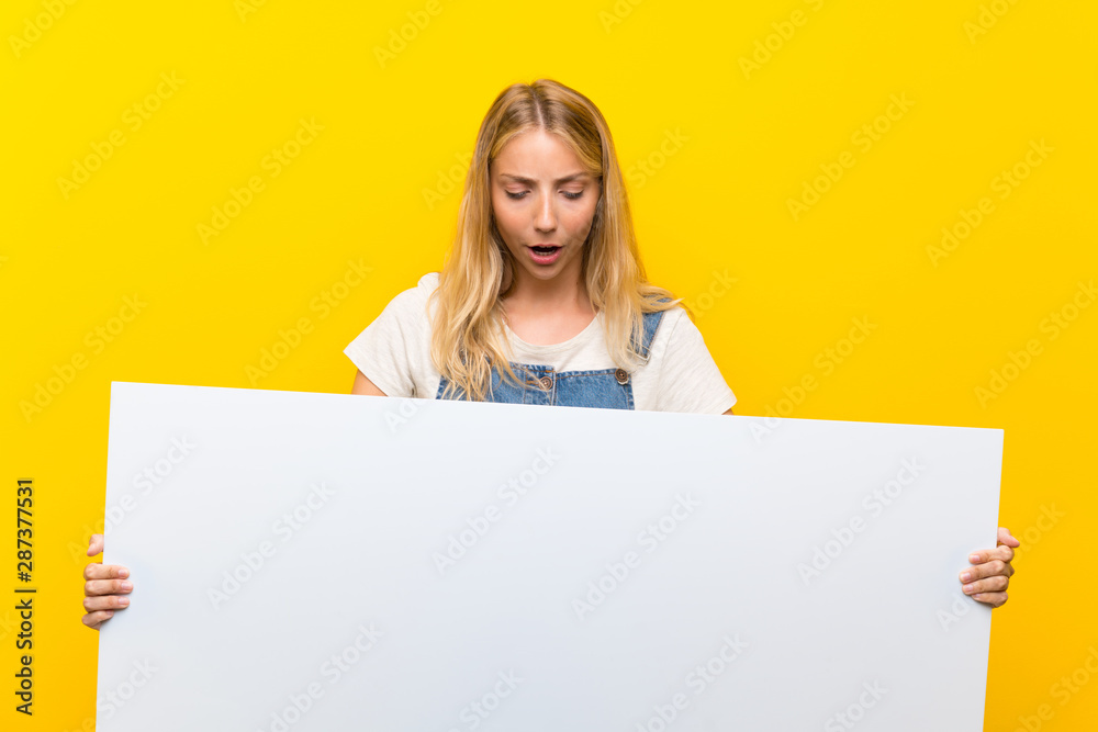Blonde young woman over isolated yellow background holding an empty white placard for insert a concept Blonde young woman over isolated yellow background holding an empty white placard for insert a concept