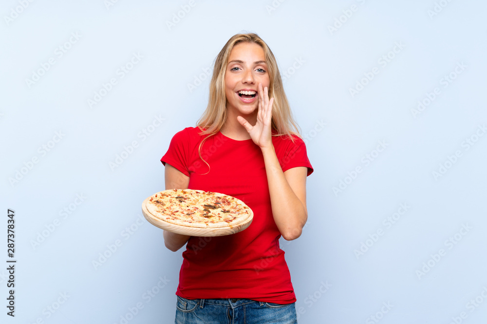 Young blonde woman holding a pizza over isolated blue wall with surprise and shocked facial expression Young blonde woman holding a pizza over isolated blue wall with surprise and shocked facial expression