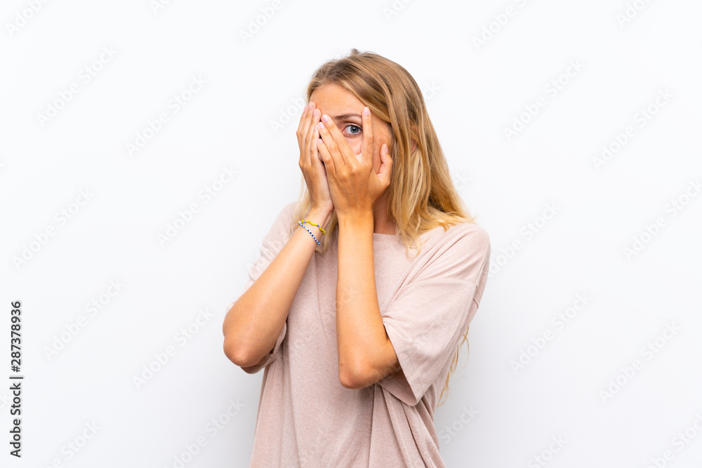 Blonde young woman over isolated white background covering eyes and looking through fingers Blonde young woman over isolated white background covering eyes and looking through fingers