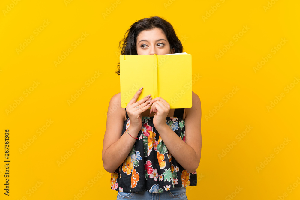 Young woman over isolated yellow background holding and reading a book - obrazy, fototapety, plakaty 