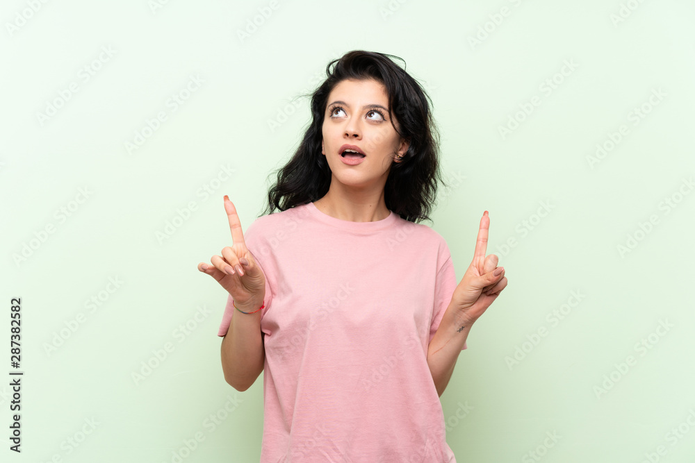 Young woman over isolated green background surprised and pointing up Young woman over isolated green background surprised and pointing up