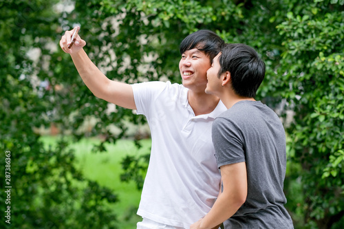 Asian gay couple take photo selfies of themselves together at balcony with green tree background and day light.