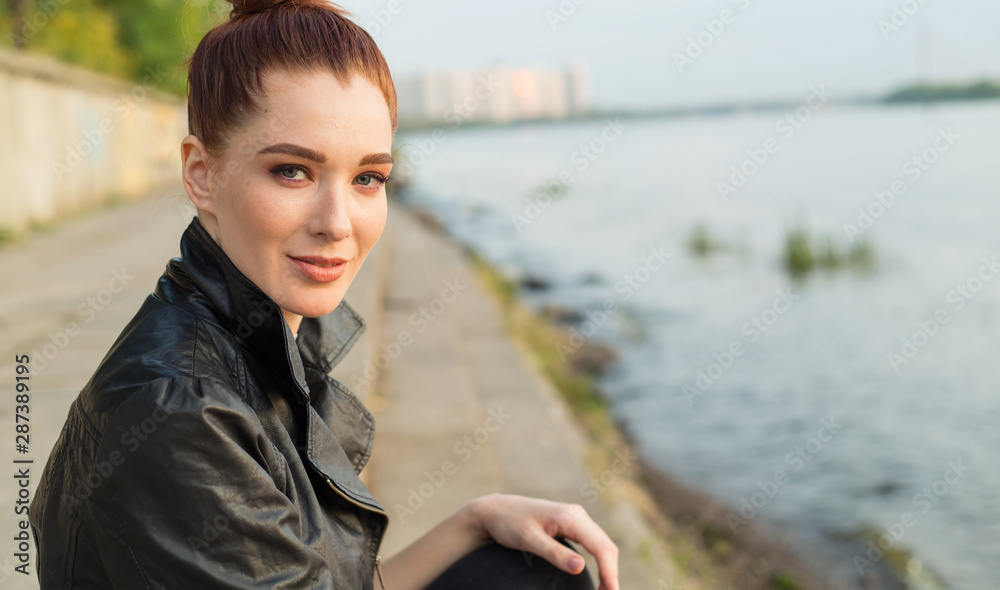 Close up portrait. Beautiful smiling red haired woman in a leather jacket sits on the river embankment. Close up portrait. Beautiful smiling red haired woman in a leather jacket sits on the river embankment.