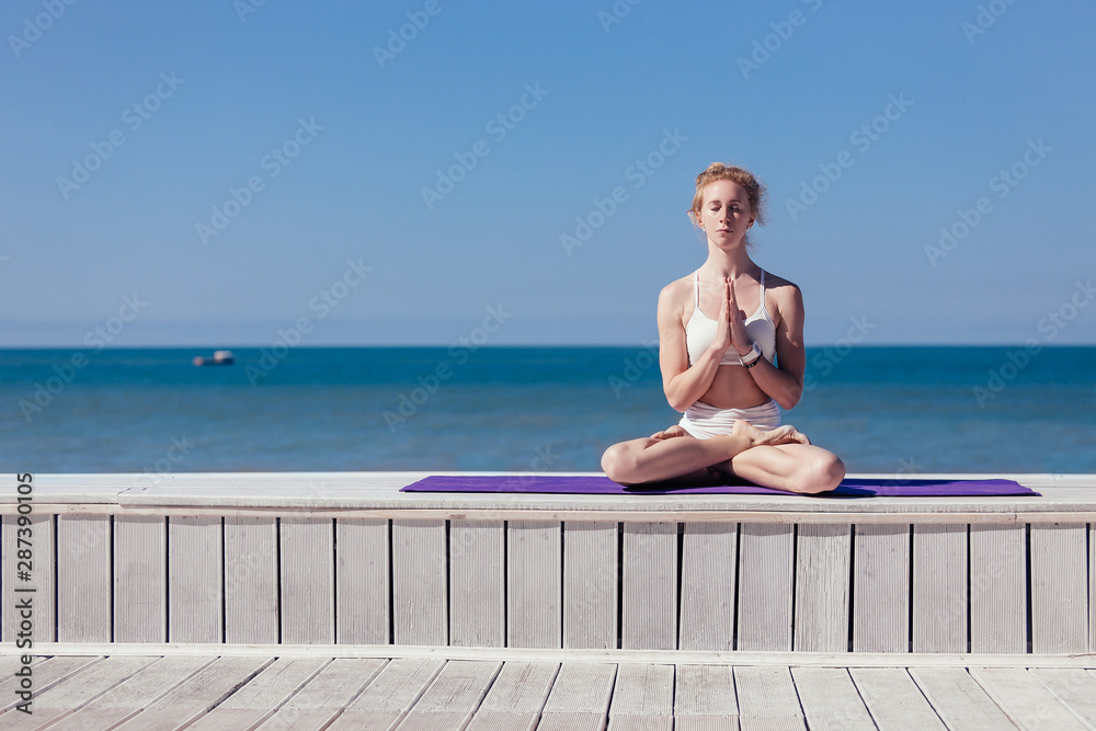 Sporty young woman sitting in lotus with namaste gesture, meditating exercise, relaxation pose. Slim girl practicing yoga outdoor by the sea, blue sky, wooden terrace. Calm, relax, healthy concept