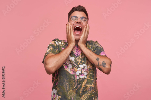 Handsome young short haired male in flowered t-shirt standing over pink background with raised palms to his face, heehawing with thrown back head photo