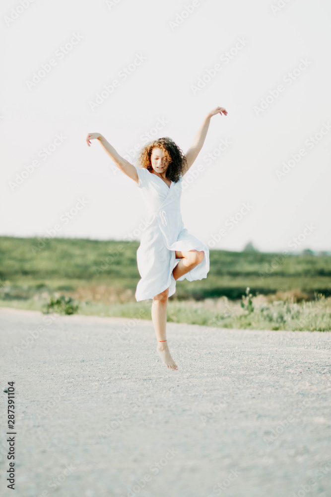 curly young woman dancing romantically in a blue dress on a sandy road at sunset. curly young woman dancing romantically in a blue dress on a sandy road at sunset.