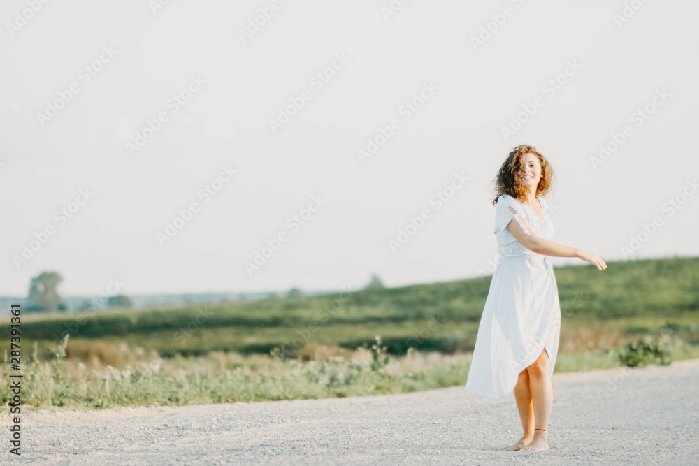 curly young woman dancing romantically in a blue dress on a sandy road at sunset. curly young woman dancing romantically in a blue dress on a sandy road at sunset.