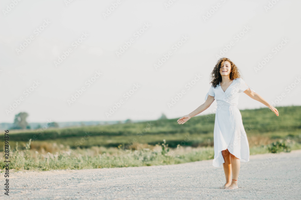 curly young woman dancing romantically in a blue dress on a sandy road at sunset. curly young woman dancing romantically in a blue dress on a sandy road at sunset.