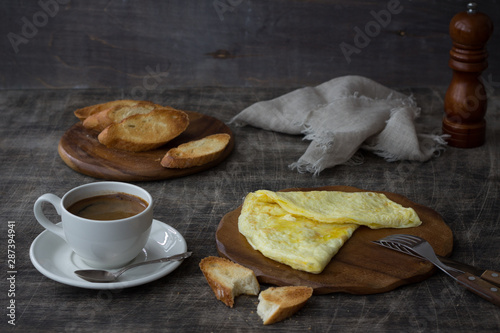 Morning breakfast of a traditional French omelet with toasts and butter and a white porcelain cup of coffee on a wooden board placed on a dark wooden table and dark wooden background