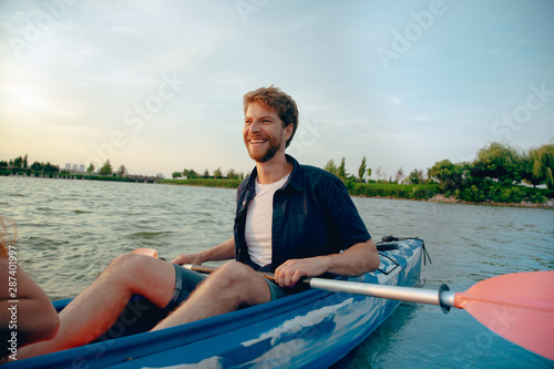 Confident young caucasian man kayaking on river with sunset in the backgrounds. Having fun in leisure activity. Happy male model laughting on the kayak. Sport, relations concept.