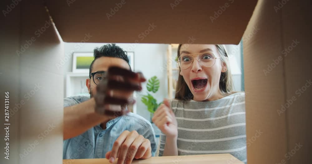 Slow motion of surprised girl and guy opening carton box taking gift laughing having fun together indoors at home. Youth, people and surprises concept.