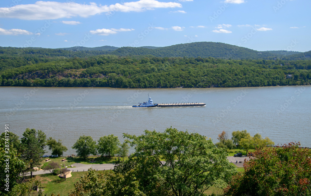 Blue and white tug boat was seen pushing a long barge down the Hudson River. There are small green tree covered hills in the background