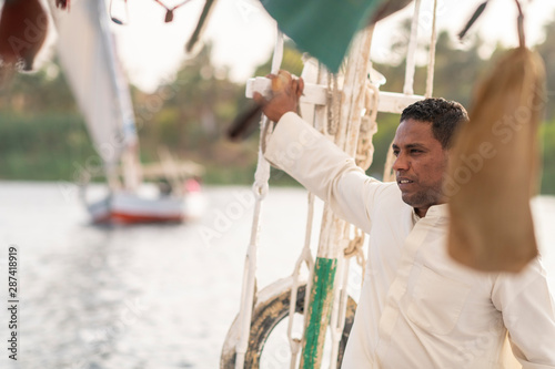 An Egyptian man holiding the mast while sailng a traditional Felucca sailboat with wooden masts and cotton sails on the Nile, Aswan, Egypt photo