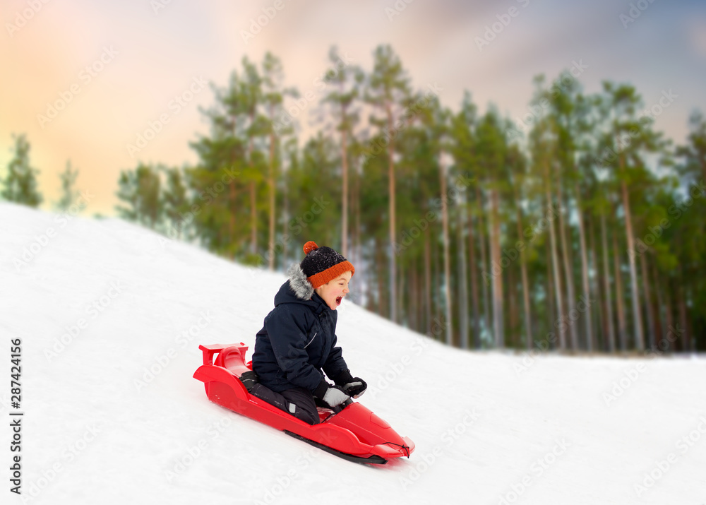 childhood, sledging and season concept - happy little boy sliding on sled down snow hill outdoors in winter over forest background