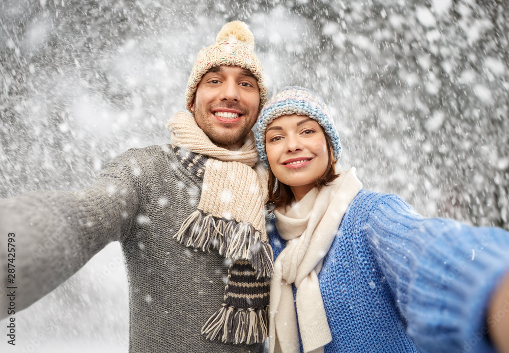 people, christmas and winter clothes concept - happy couple in knitted hats and scarves taking selfie over snow background