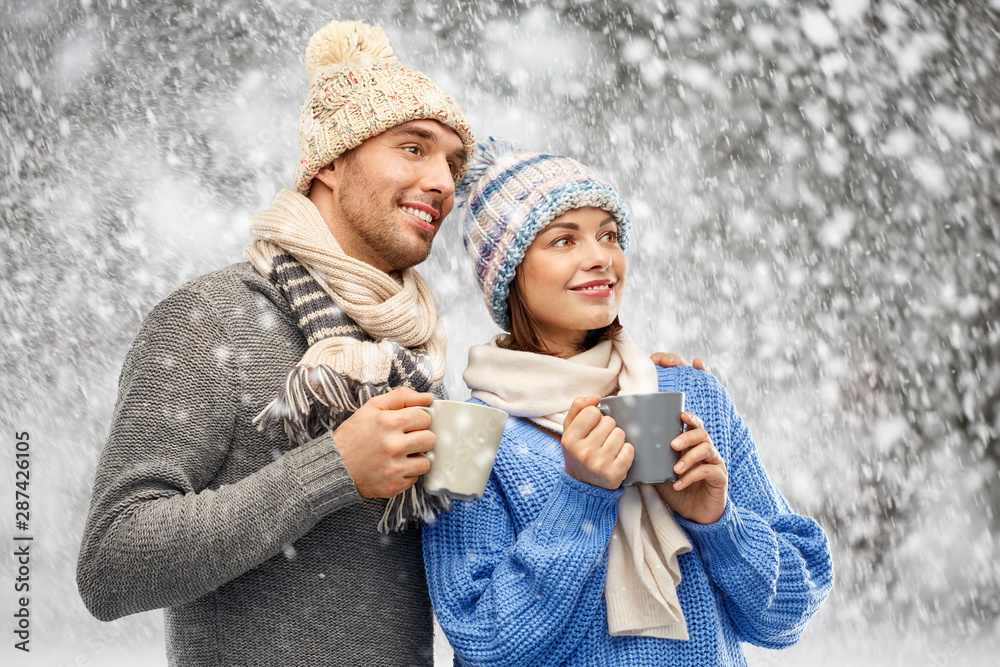 Fototapeta premium people, christmas and love concept - happy romantic couple in knitted hats and scarves with mugs over snow background