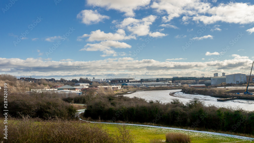 Photo taken from the Northern Spire Bridge with the river Wear flowing towards Sunderland and the sea. Also showing embankments either side, the Queen Alexandra Bridge and buildings in the distance.