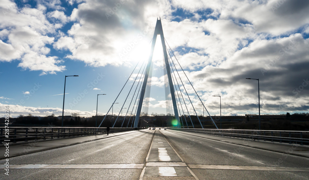 Northern Spire Bridge (opened Summer 2018) in Sunderland spanning the River Wear.  Photo taken facing the sun giving the bridge a silhouette contrasting with the blue sky with dark and white clouds.