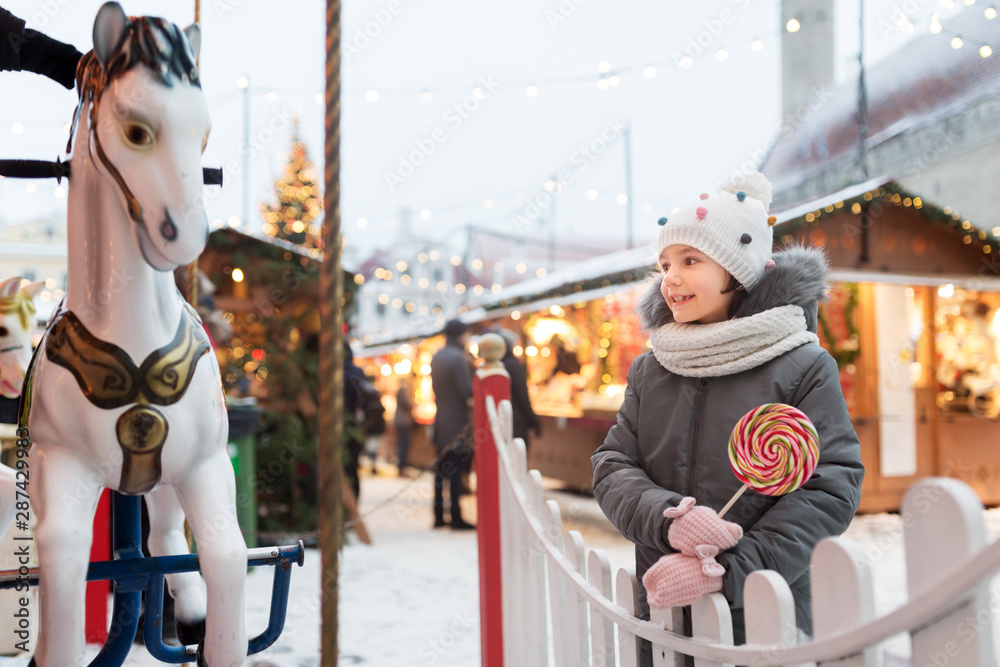 holidays, childhood and people concept - happy little girl with big lollipop at christmas market