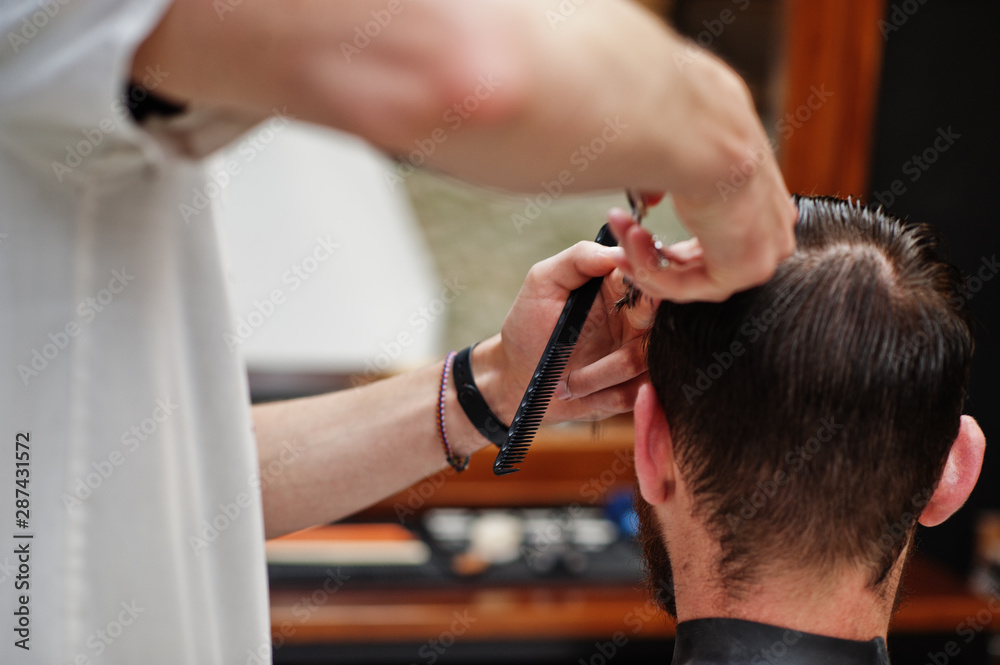 Young bearded man getting haircut by hairdresser while sitting in chair at barbershop. Barber soul.