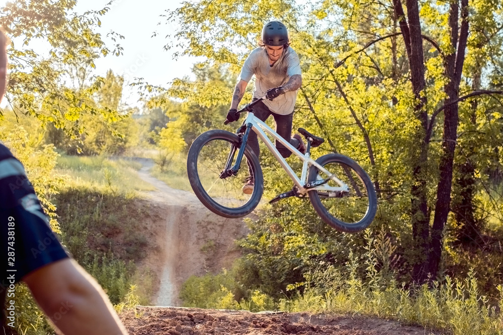 Obraz premium a cyclist watches his partner jumping on a bicycle and performing tricks. in the forest against the setting sun