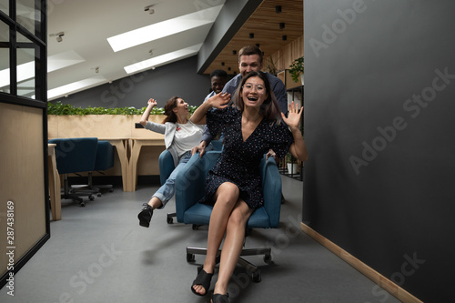Happy funny diverse coworkers riding on chairs in modern office