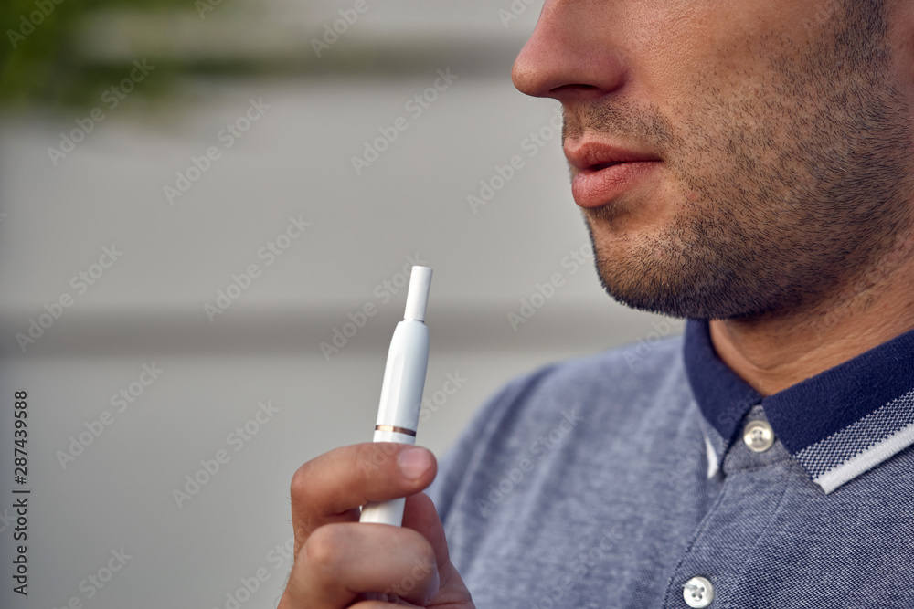 Electronic cigarettes, cigarette technology. Tobacco IQOS system. Close-up of a man smoking an electric hybrid cigarette with a heatstick. New tobacco heating system without burning. Electronic cigarettes, cigarette technology. Tobacco IQOS system. Close-up of a man smoking an electric hybrid cigarette with a heatstick. New tobacco heating system without burning.