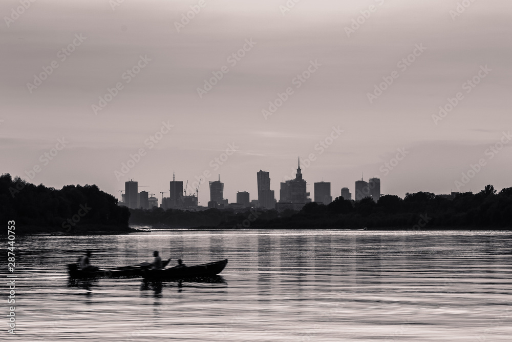 Monochrome Warsaw cityscape with skyscrapers in the downtowan and two boats sailing on the Vistula River during sunset.