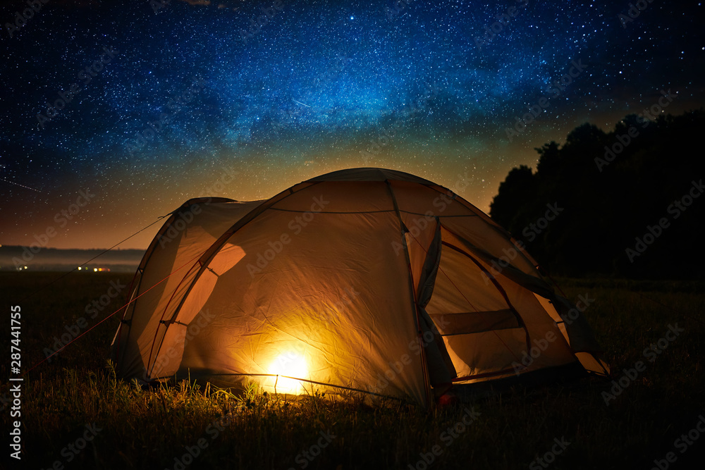 Traveling and camping concept - camp tent at night under a sky full of stars. Orange illuminated tent. Beautiful nature - field, forest, plain. Moon and moonlight