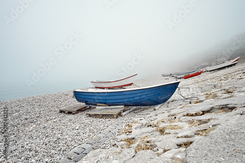 Εκτύπωση καμβά Blue and red fishing boats on a rocky pebble beach in the early morning with fog