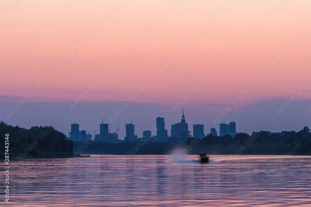 Colorful Warsaw cityscape with skyscrapers in the downtowan and one boat sailing fast on the Vistula River during sunset.