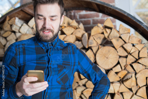 Smiling bearded hipster guy reading text message in social network via mobile phone standing against firewood design background with copy space. Cheerful businessman chatting on cell telephone