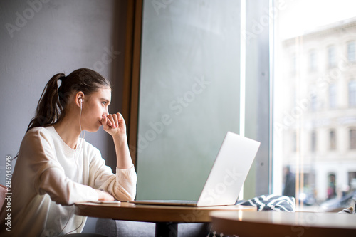 Sad hipster girl thinking about something listening to music via headphones and looking in window while sitting with laptop computer in coffee shop. Thoughtful woman dreaming after work on notebook