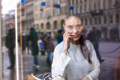 View through coffee shop window with street reflection of a hipster girl having mobile phone conversation with friend while sitting in restaurant. Woman talking on cell telephone during rest in cafe