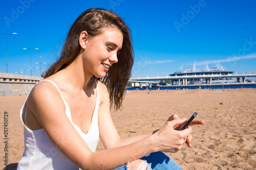 Happy smiling woman chatting online with friends via mobile phone while relaxing outdoors on the beach in sunny summer day. Cheerful female watching video on cell telephone, resting on seashore