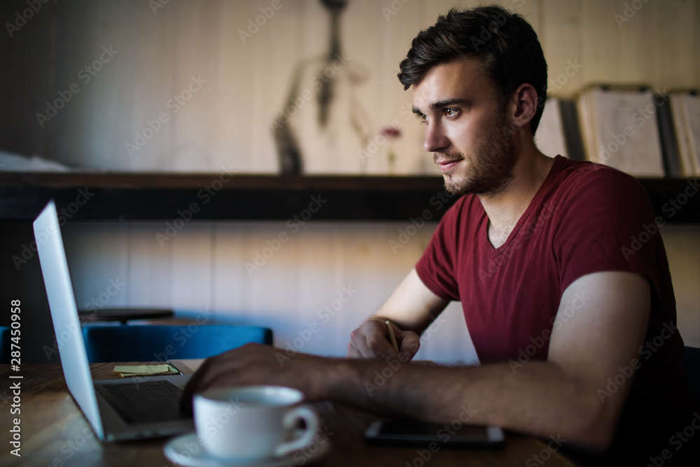 Handsome hipster guy skilled freelancer distance working on laptop computer while sitting in coffee shop during free time. Male successful copywriter preparing an article for publication using netbook Handsome hipster guy skilled freelancer distance working on laptop computer while sitting in coffee shop during free time. Male successful copywriter preparing an article for publication using netbook