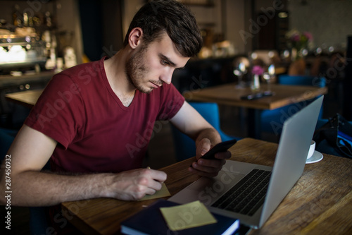 Young serious man writes the information on the sticker from e-mail on mobile phone during webinar on laptop computer. Hipster guy skilled freelancer writing notary during online training course