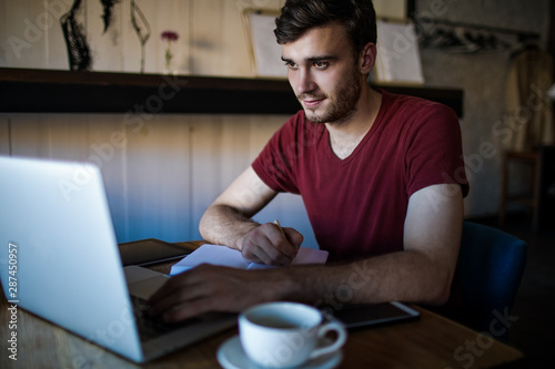 Man college student writing notary in notepad during online education on laptop computer, sitting with cup of coffee in modern restaurant. Handsome male skilled content writer working on web site