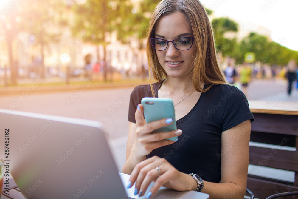 Gorgeous woman in glasses with beautiful smile on the face reading pleasant text message in messenger via smartphone during online learning on laptop computer, sitting in coffee shop near copy space Gorgeous woman in glasses with beautiful smile on the face reading pleasant text message in messenger via smartphone during online learning on laptop computer, sitting in coffee shop near copy space