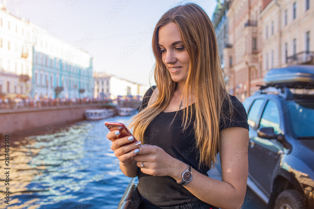 Fototapeta premium Young gorgeous woman online ordering via mobile phone while standing outdoors near Neva river background with copy space for promotional content. Pretty female traveler booking via cellphone