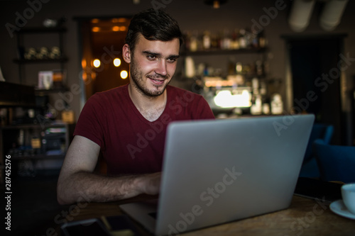 Smiling man freelance social media content writer reading article on web site via laptop computer while sitting in modern restaurant. Male marketing coordinator having video conference via netbook