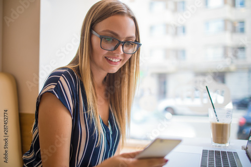 Happy smiling woman freelance social media content writer reading text message on mobile phone during work on laptop computer,sitting in coffee shop during recreation time.Joyful hipster girl chatting