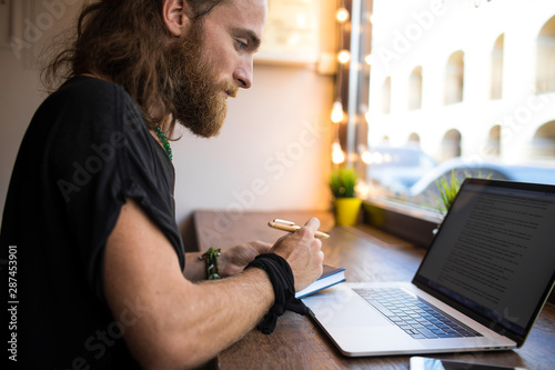 Serious bearded Sweden man with long hair having online training course via laptop computer, writing notary while sitting in modern coworking space. Male project manager working distance on netbook