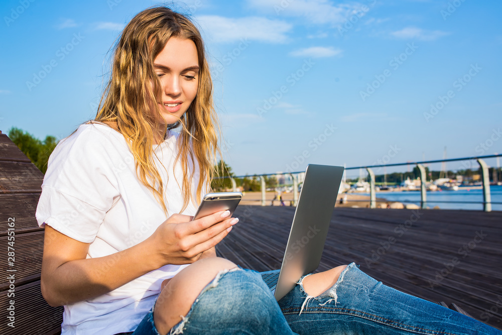 Beautiful smiling Caucasian woman reading lifestyle blog on smartphone during online education on portable laptop computer while sitting outdoors in sunny summer day. Female chatting on cell telephone