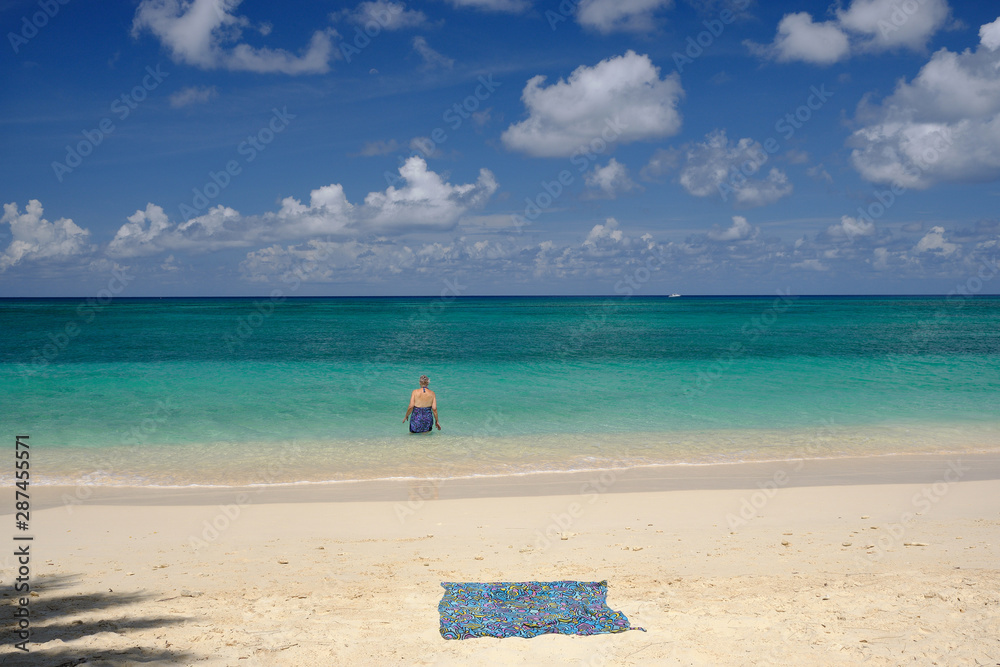 A silver gray haired woman enters the crystal clear waters of the Cayman Islands on a deserted beach leaving a beach towel in the sand A silver gray haired woman enters the crystal clear waters of the Cayman Islands on a deserted beach leaving a beach towel in the sand
