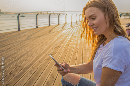 Young cheerful woman reading notifications on mobile phone while sitting outdoors on wooden embankment background with copy space for advertising in sunny summer evening. Female chatting on smartphone